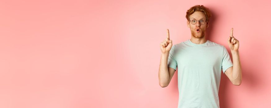 Impressed caucasian man with ginger hair, wearing glasses and t-shirt, saying wow and pointing fingers up at awesome deal, standing over pink background.