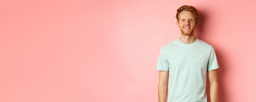 Happy redhead man in t-shirt looking at camera and smiling, standing over pink background.