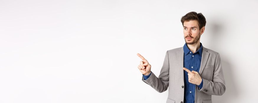 Handsome businessman in suit pointing, looking left at logo with serious face, standing against white background.