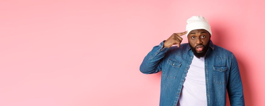 Annoyed and pissed-off african-american man pointing finger at head, scolding someone stupid, staring bothered at camera, pink background.