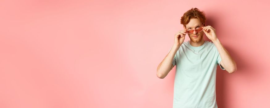 Handsome redhead man in summer sunglasses looking sassy at camera, standing over pink background.