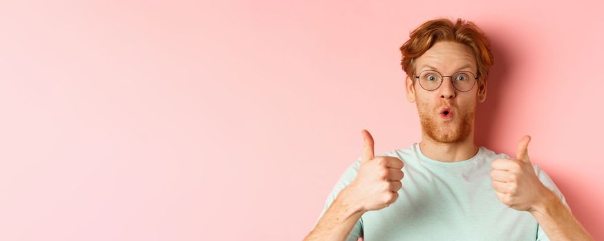 Face of happy redhead man in glasses and t-shirt, showing thumbs-up and looking excited, approve and praise cool promotion, standing over pink background.