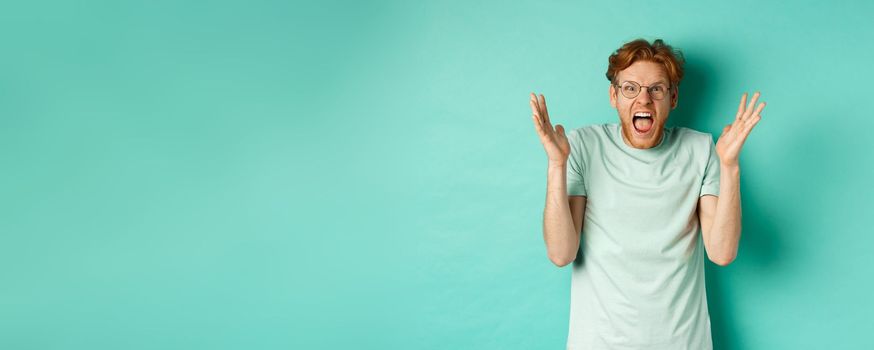 Portrait of distressed and angry redhead guy losing temper, shouting and shaking hands outraged, staring with furious face at camera, standing over mint background.