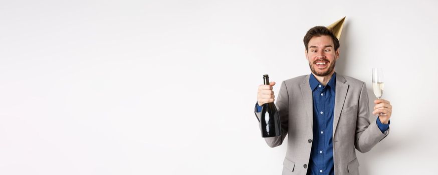 Celebration and holidays concept. Funny drunk guy in suit and birthday hat, squinting eyes and having fun at party, drinking champagne, white background.