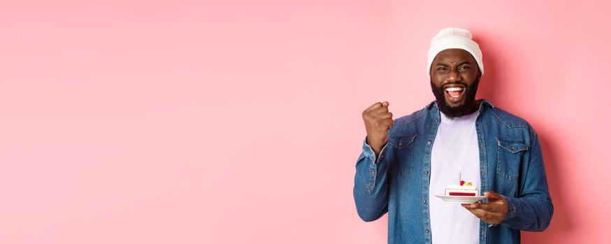 Cheerful african-american guy celebrating birthday, making wish on bday cake with lit candle, smiling happy, standing over pink background.