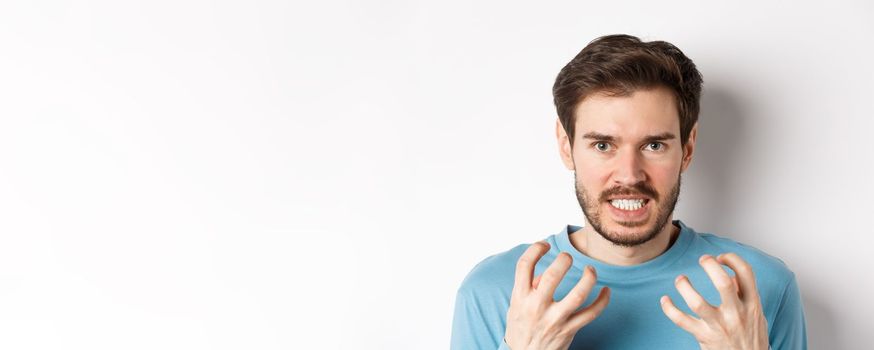 Close up of angry young man with beard, shaking hands mad, squeeze teeth and frowning furious, standing outraged over white background.