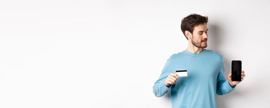 Mobile banking. Handsome caucasian man showing plastic credit card and looking at blank mobile screen, standing over white background.