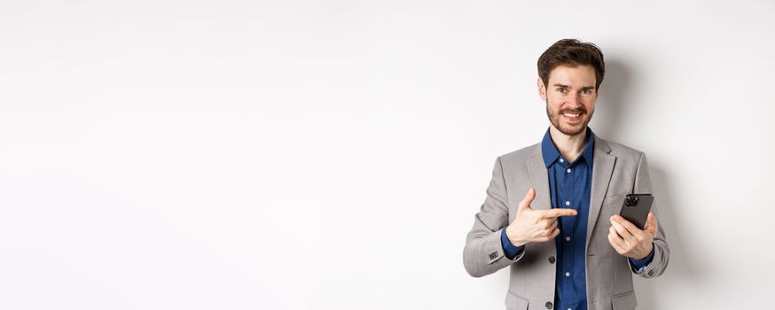 Happy successful man in suit pointing at cellphone, advertising mobile app, standing on white background.
