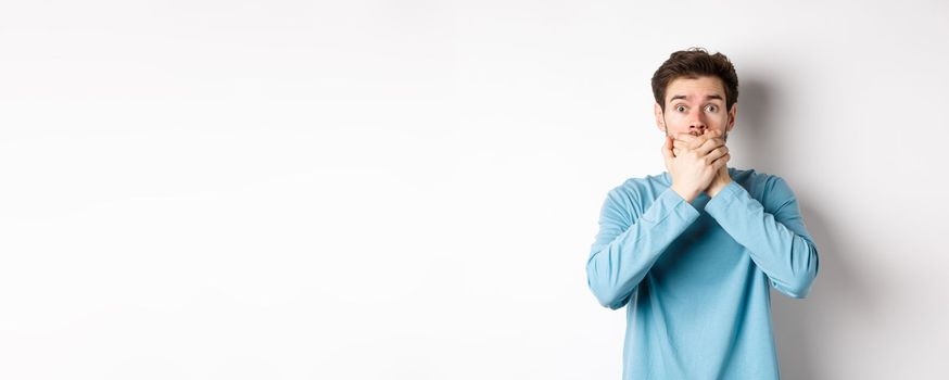 Image of shocked young man staring startled and covering mouth with hands, hear gossips, standing on white background.