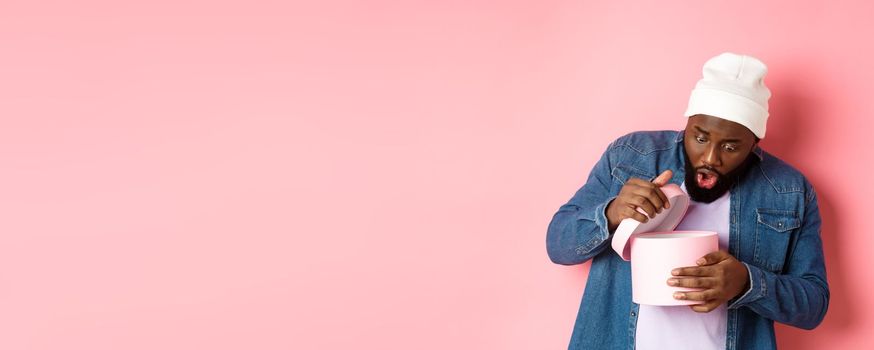 Image of surprised african-american man open box with birthday gift, staring at present with amazement, standing against pink background.