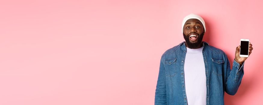 Handsome hipster guy in beanie and denim shirt smiling, showing mobile phone screen with happy face, recommend app, standing over pink background.