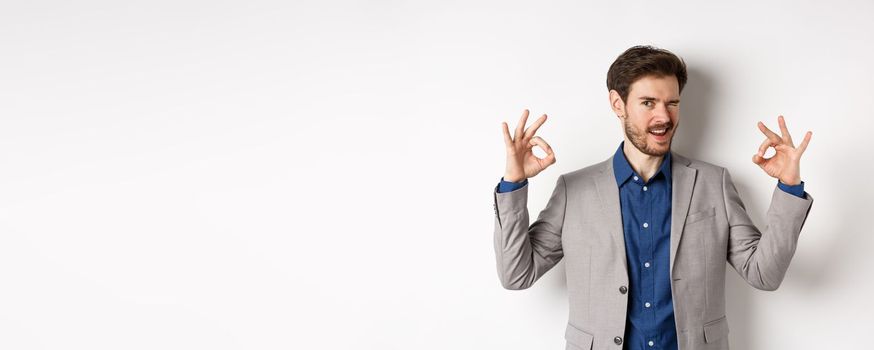 Cheeky handsome man in suit winking at you, showing okay signs, all under control gesture, approve good choice, assuring everything OK, standing on white background.