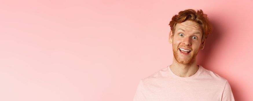 Headshot of funny redhead guy showing tongue, making silly faces at camera, standing joyful against pink background. Copy space