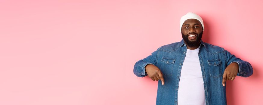 Handsome hipster african-american man pointing fingers down, smiling and showing promo offer, standing over pink background.