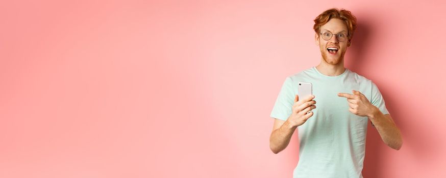 Cheerful guy talking about internet promo, smiling amazed and pointing finger at smartphone, standing over pink background.
