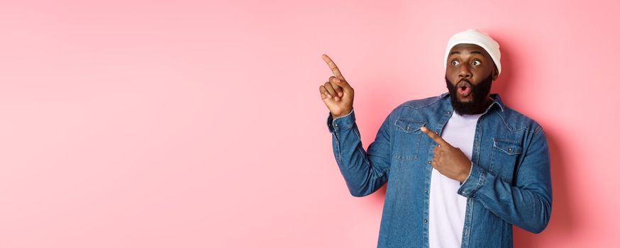 Impressed hipster black man in beanie and denim shirt, looking and pointing at upper left corner logo, staring amazed, standing over pink background.