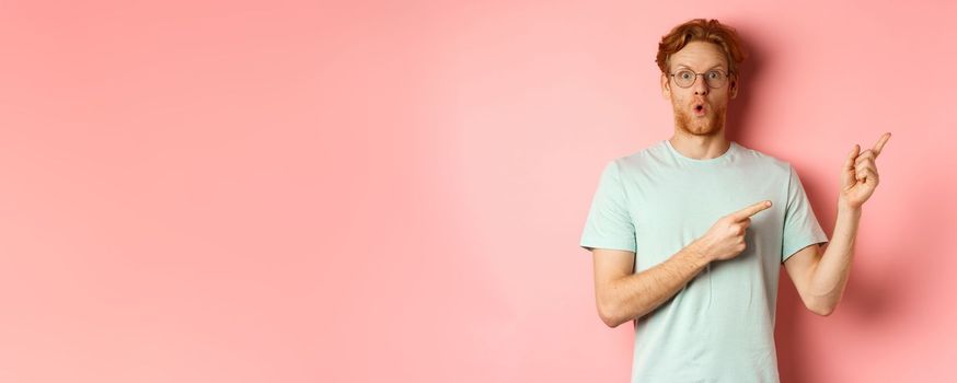 Impressed caucasian guy with red beard and hair, pointing fingers at upper right corner and gasping amazed, saying WOW, standing over pink background.