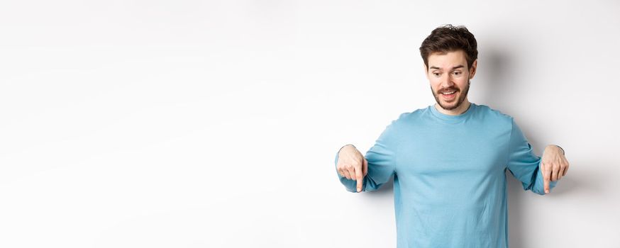 Excited and happy young man looking and pointing down at awesome deal, checking out promotion offer, standing on white background.