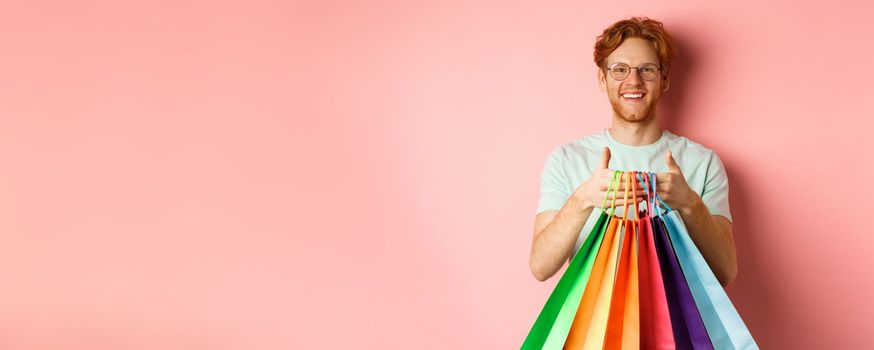 Cheerful redhead man buying gifts, holding shopping bags and smiling, standing over pink background.