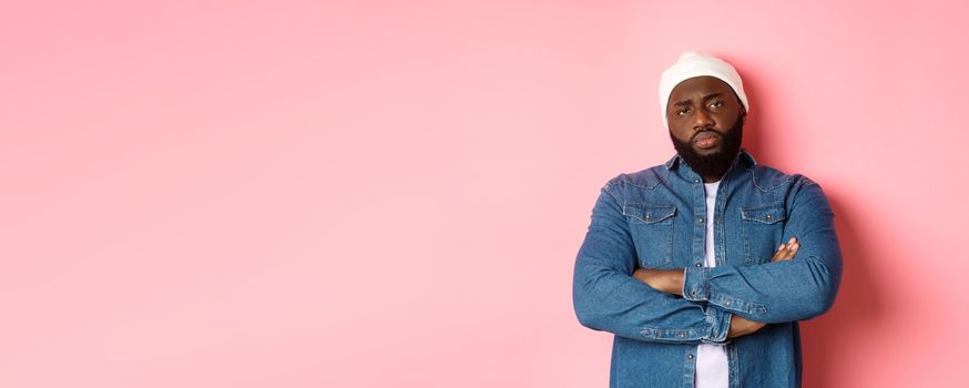 Disappointed african-american man in hipster beanie, staring at camera displeased, cross arms on chest, standing over pink background.