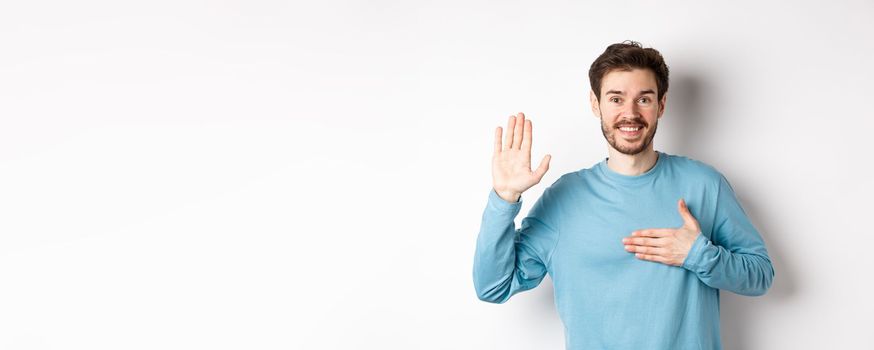 Smiling hosent man raising arm and hold hand on heart, making promise tell truth, swear or give oath, standing over white background.