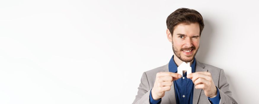 Real estate and insurance concept. Handsome man in suit winking and smiling, showing small paper house cutout, standing on white background.