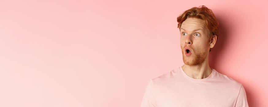 Close up of shocked redhead man with beard, saying wow, looking left with amazed face, standing over pink background.