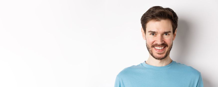 Close-up of handsome caucasian man smiling with white teeth, looking confident at camera, standing in blue shirt on white background.