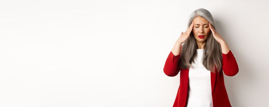 Tired senior businesswoman in red blazer having headache, touching head and feel sick, standing with migraine against white background.