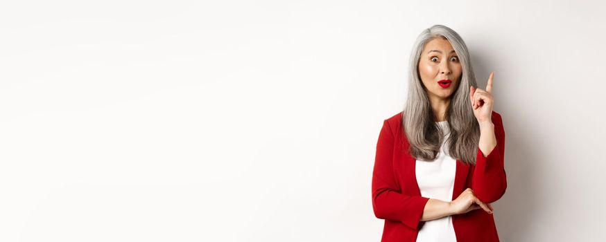 Elderly asian woman entrepreneur in red blazer having an idea, suggesting something, raising finger in eureka gesture, standing over white background.