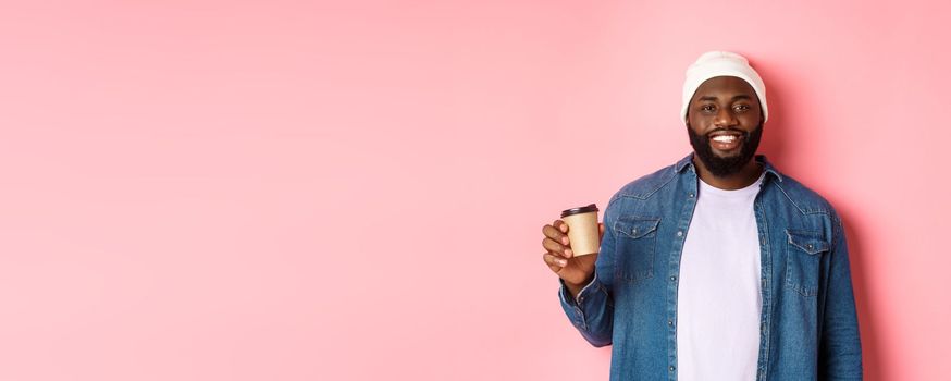 Handsome modern Black man drinking takeaway coffee, smiling and looking satisfied at camera, standing over pink background.