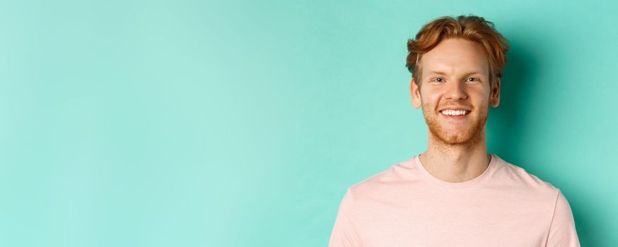 Close up of redhead bearded guy in pink t-shirt, smiling with white perfect teeth and looking at camera, standing over turquoise background.