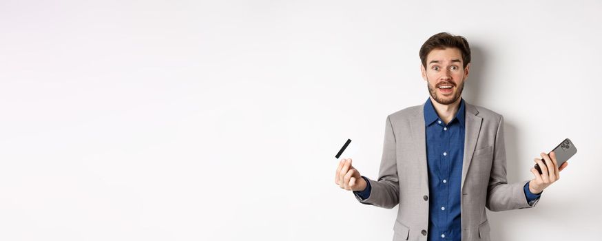 Online shopping. Surprised businessman holding plastic credit card and smartphone, making money in internet, standing in suit against white background.