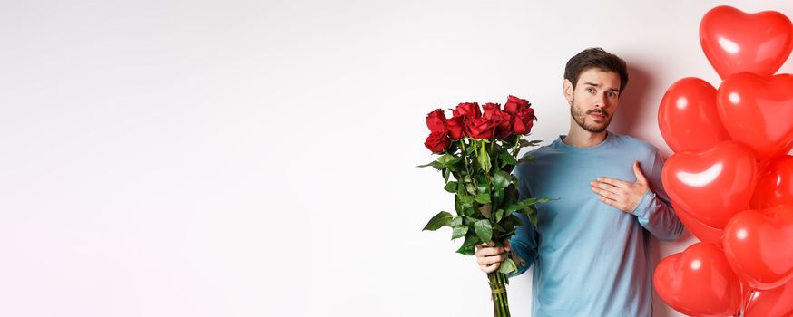 Romantic guy express his love on Valentines day with gifts, bring bouquet of red roses and balloons, holding hand on heart, standing over white background.