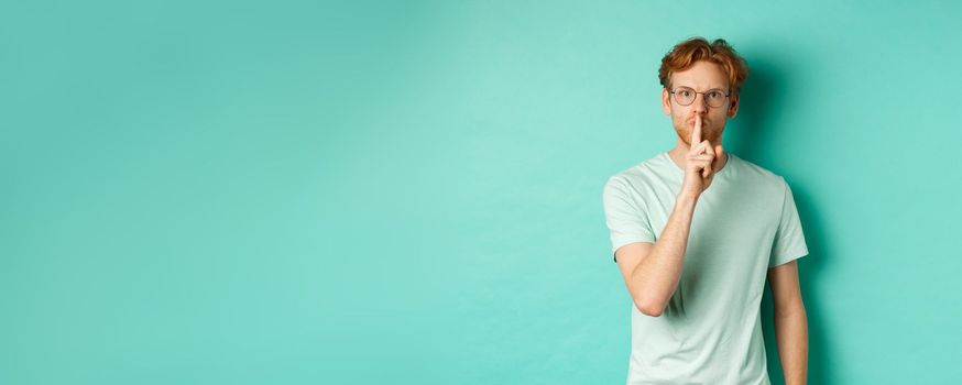 Serious redhead man hushing, tell to be quiet, making shush gesture and looking at camera, standing in t-shirt over turquoise background.