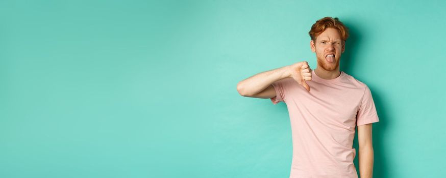 Disappointed redhead man showing thumbs-down and tongue, epxress dislike and disapproval, standing over turquoise background.