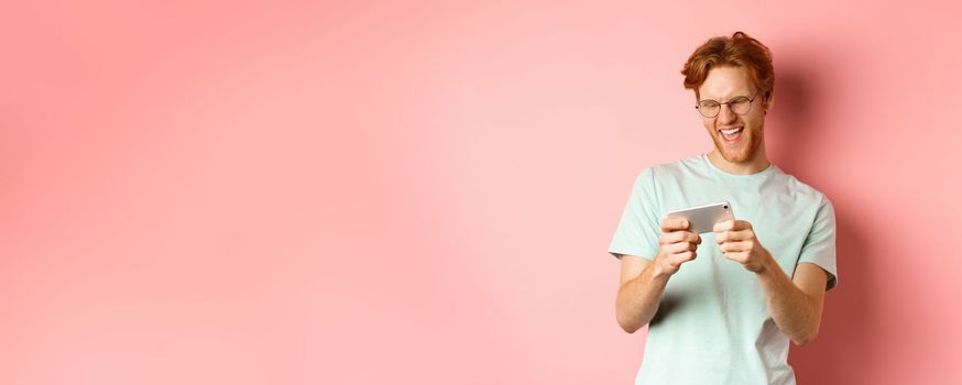 Happy young man with red messy haircut, wearing glasses, playing video game on smartphone and having fun, looking at mobile screen, standing over pink background.