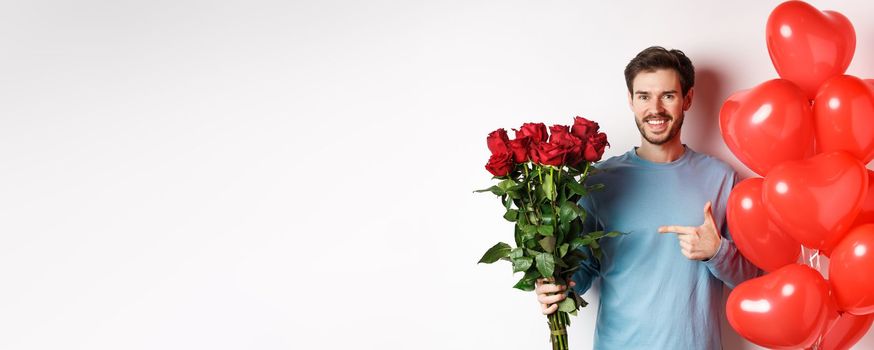 Valentines day romance. Happy young man with flowers and heart balloons going on date with lover. Smiling guy pointing at bouquet of red roses, standing over white background.