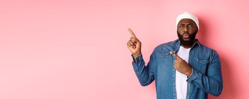 Confused and displeased african american man grimacing, cringe from disappointment and pointing fingers at upper left corner, bad promo, pink background.