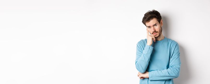 Skeptical young man listening to you with bored face, looking at camera reluctant, standing in sweatshirt over white background.