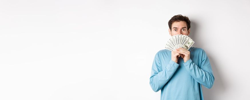 Surprised and amazed handsome guy showing money, looking at promo offer, going shopping with cash, standing over white background.