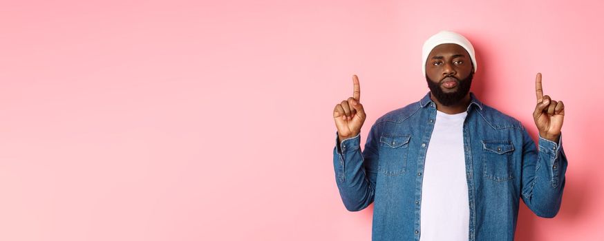 Serious and bothered Black man with beard, staring at camera and pointing fingers up, showing promo, standing over pink background.