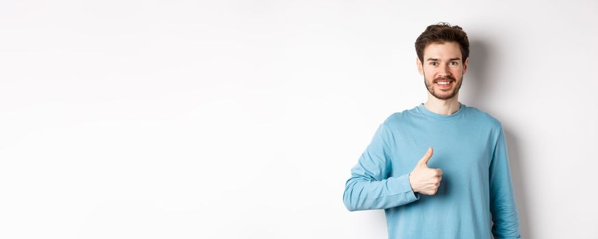 Smiling male model in casual shirt showing thumb up, approve and recommend product, praise excellent choice, standing on white background.