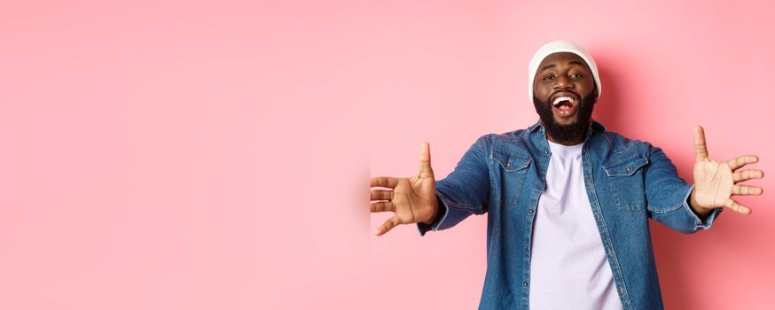 Happy Black man stretching hands, reaching for hug or take something, smiling pleased, standing in beanie and denim shirt over pink background.