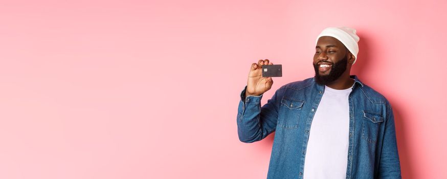 Shopping concept. Image of happy african-american man looking satisfied at credit card, recommending bank, standing over pink background.