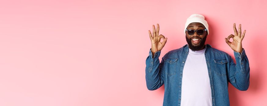 Image of cool african-american hipster man showing okay signs in approval, like something awesome, wearing sunglasses and beanie, pink background.