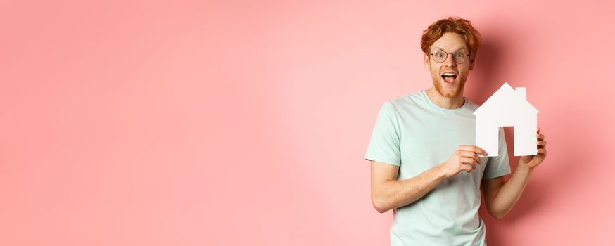 Real estate. Cheerful redhead man buying property, showing paper house cutout and smiling amazed, standing over pink background.