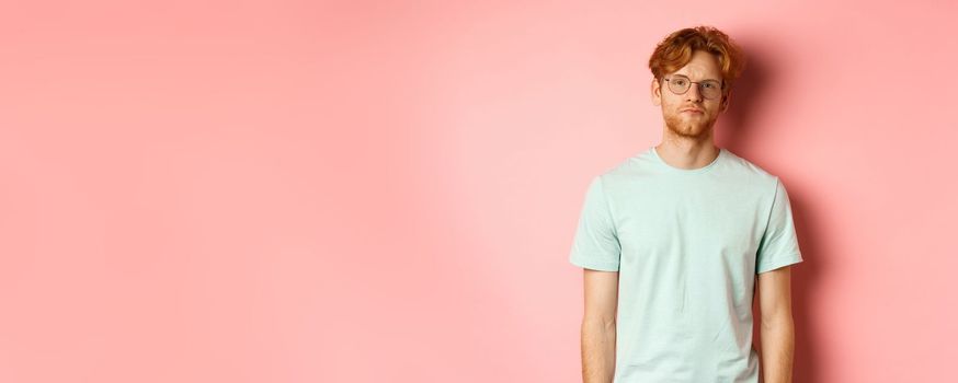 Sad and gloomy redhead bearded man in t-shirt and glasses, staring at camera bored and unamused, standing over pink background.