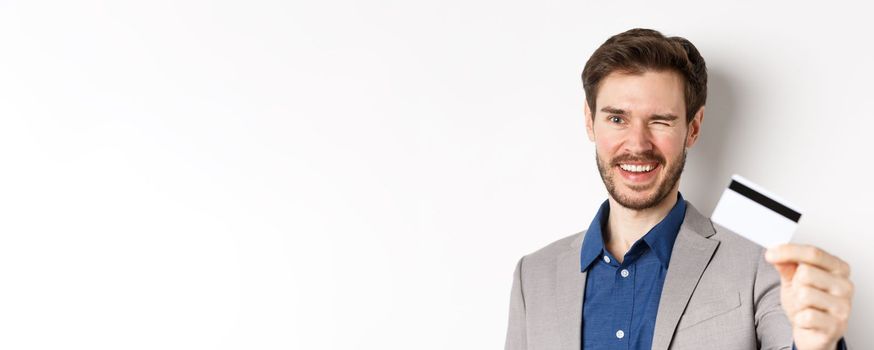 Shopping. Cheerful bearded guy in suit winking and smiling, showing plastic credit card at camera, white background.