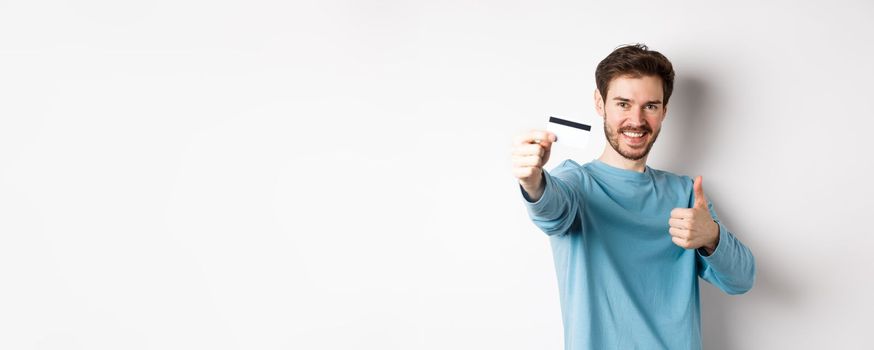 Handsome caucasian man showing plastic credit card with thumb-up, recommend and praise good bank offer, standing on white background